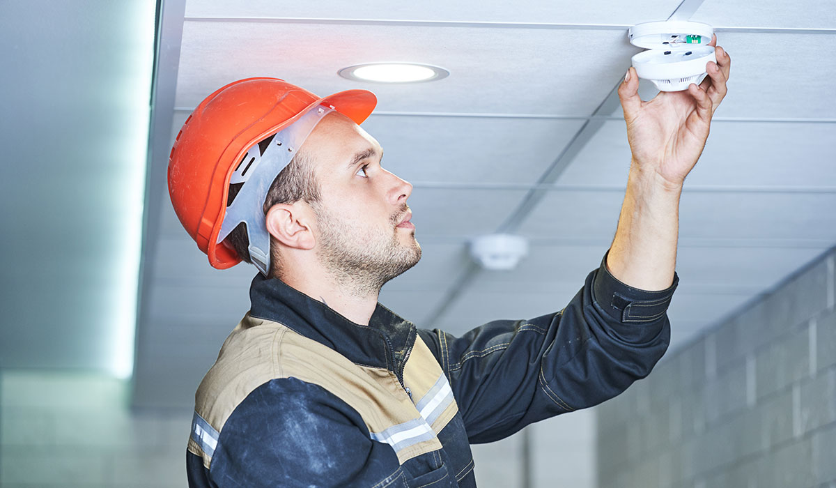 A person in a hardhat inspecting a ceiling smoke detector.